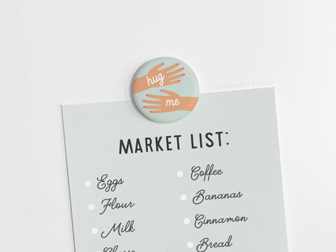 The Hug Me Hands Button Magnet by Onderkast Studio features playful illustrated arms hugging a “Market List” with eggs, flour, milk, coffee, bananas, cinnamon, and bread—a delightful reminder for your next shopping trip.