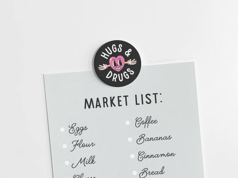 A market list with items like eggs, flour, and milk is held to a white surface by the Onderkast Studio Hugs & Drugs Button Magnet, featuring a smiling pink heart with open arms and the text Hugs & Drugs.