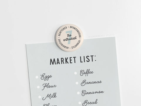 A light blue market list featuring eggs, flour, milk, coffee, bananas, and bread is held by the Onderkast Studio Enneagram Button Magnet – 7 The Enthusiast, perfect for celebrating the creative spirit of Type 7s.
