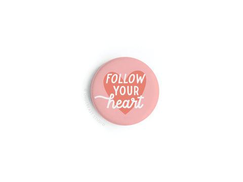 The Follow Your Heart Deep Button Magnet by Onderkast Studio displays FOLLOW YOUR heart in white and orange lettering over a heart shape, all set against a crisp white background with vibrant, round pink design.
