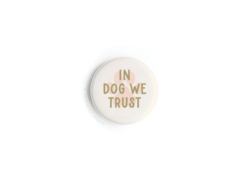 The In Dog We Trust Button Magnet by Onderkast Studio features bold gold lettering on a white round magnet, accented with a pastel dog’s nose and ears—an ideal tribute for canine lovers.