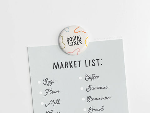 A round Social Loner Button Magnet by Onderkast Studio, printed in the USA, secures a blue market list—featuring eggs, flour, milk, cheese, coffee, bananas, cinnamon, and bread—on a white surface with its eye-catching design.