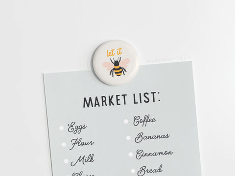The Onderkast Studio Let It Bee Button Magnet, featuring a colorful bee illustration, securely holds a market list with eggs, flour, milk, coffee, bananas, cinnamon, and bread on a light surface.