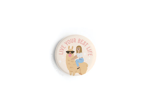 The Live Your Best Life Button Magnet by Onderkast Studio showcases a cartoon girl riding a sunglasses-wearing llama, vibrant colors, and the text LIVE YOUR BEST LIFE on a light pink background.