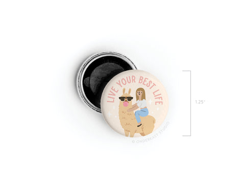 The Live Your Best Life Button Magnet by Onderkast Studio is a 1.25 round magnet featuring colorful print, a llama in sunglasses with a smiling woman riding on its back, and the phrase LIVE YOUR BEST LIFE in pink text above them.
