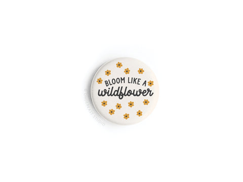 The Bloom Like a Wildflower Button Magnet by Onderkast Studio features vibrant black and cursive text with small yellow flowers on a round white background.