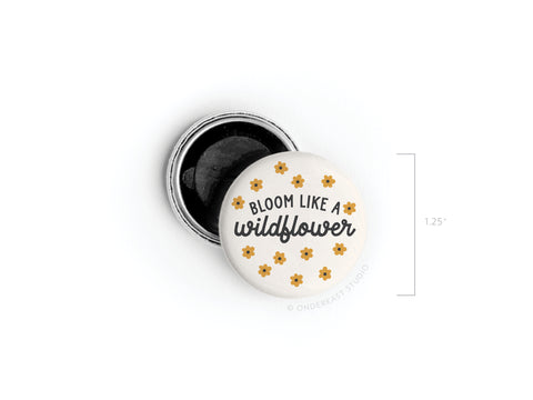 The Bloom Like a Wildflower Button Magnet by Onderkast Studio is a 1.25-inch round pin featuring yellow flowers and black text, displayed with its open backing.
