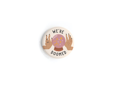 The Onderkast Studio We’re Doomed Button Magnet features two hands with painted nails holding a sparkly pink crystal ball above “WERE DOOMED” text, all vibrantly printed on a white background.