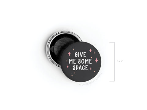 A round black tin from Onderkast Studio holds a black balm inside. Its lid, embossed with GIVE ME SOME SPACE in white and colorful stars, is off. A 1.25-inch measurement appears next to the Give Me Some Space Button Magnet.