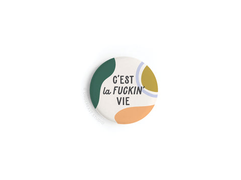 The C’est La Fuckin’ Vie Button Magnet by Onderkast Studio features bold text on a white background with abstract green, yellow, blue, and orange shapes, adding playful French flair to any surface.