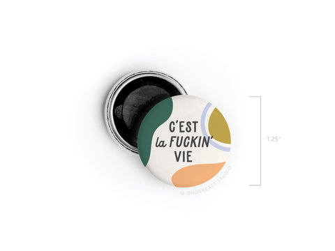 The C’est La Fuckin’ Vie Button Magnet by Onderkast Studio features colorful abstract shapes and bold text with French flair. Approx. 1.25, it comes with an open metal pin back for easy attachment.