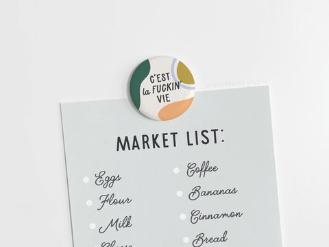 The C’est La Fuckin’ Vie Button Magnet by Onderkast Studio adds French flair atop a light blue market list with eggs, flour, milk, coffee, bananas, cinnamon, and bread.