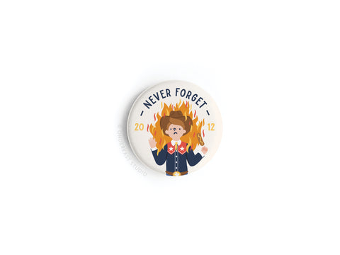 The Never Forget Button Magnet by Onderkast Studio features a shocked cartoon scout before flames with NEVER FORGET and 2012 above—ideal for fans of quirky regional humor.