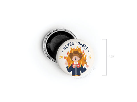The Onderkast Studio Never Forget Button Magnet features regional humor with a cartoon girl in a blue uniform and red scarf amid bright flames, framed by “NEVER FORGET” and “2012,” with the pin placed next to its open lid.