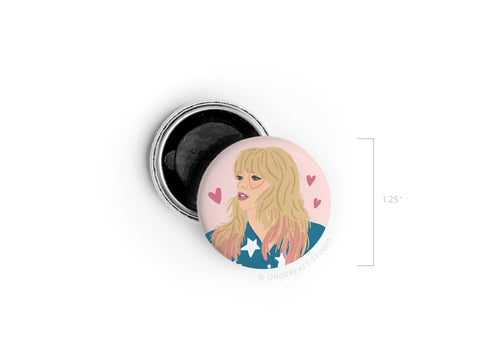 The Onderkast Studio TS Lover Button Magnet features an illustrated blonde woman with bangs in a blue and white outfit, surrounded by pink hearts. This 1.25-inch round pin includes a black back—a sweet nod to Taylor’s enchanting lyrics.