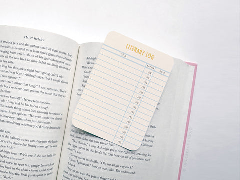 An open book displays a cream-colored Onderkast Studio Literary Log Book Review Card, made from premium card stock, with visible columns for title, rating, and date. Some text from the book appears beneath the card.