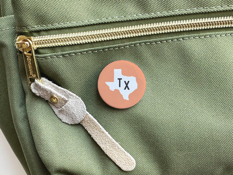 An Orange TX Pinback Button by Onderkast Studio, featuring a white outline of Texas and Tx in the center, is attached to a green fabric bag with a gold zipper and suede pull tab, adding a touch of Texan style.