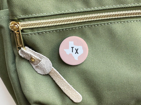 A Light Pink TX Pinback Button by Onderkast Studio is attached to the pocket of a light green fabric bag with a gold zipper and suede pull tab, adding a touch of Texas style.