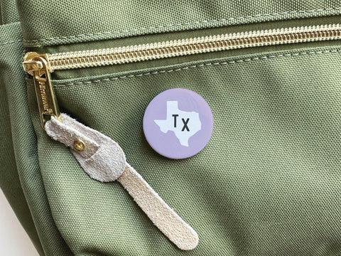 The Onderkast Studio Purple TX Pinback Button features a white Texas silhouette and TX letters, vibrant purple tones, and is perfect for showing off state pride on your favorite bag.