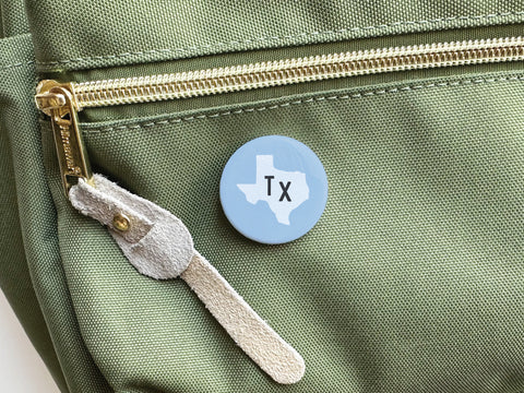 The Onderkast Studio Light Blue TX Pinback Button features a vibrant white Texas silhouette and TX letters, pinned to a green zippered bag with suede pull—perfect for showing off your state pride.