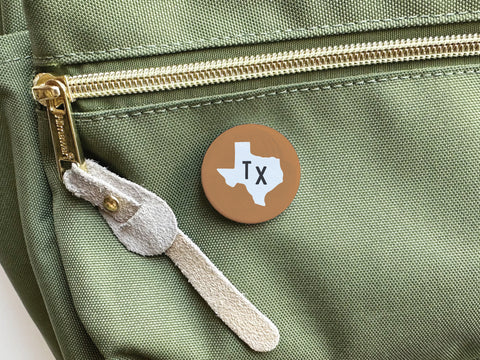 The Onderkast Studio Brown TX Pinback Button, featuring a white Texas silhouette and Tx, is attached to a green bag with a gold zipper and suede pull tab, adding a subtle touch of state pride.