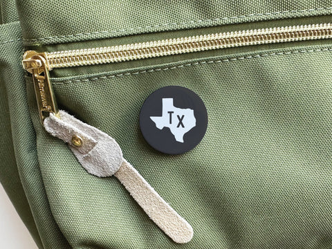 The Onderkast Studio Black TX Pinback Button, featuring a white Texas silhouette and TX letters, displays your state pride when pinned to the pocket of a green bag with a gold zipper and suede pull.