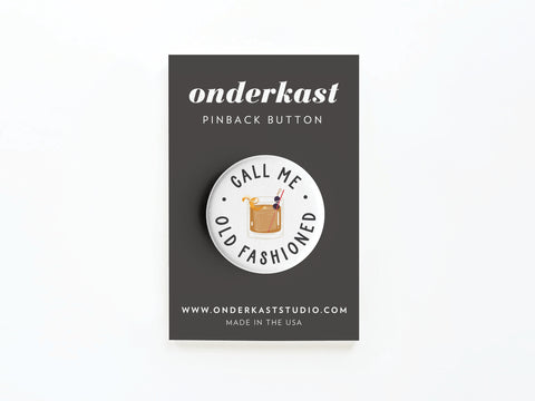The Call Me Old Fashioned Pinback Button by Onderkast Studio features a classic cocktail illustration and playful text, displayed on a dark card. Add vintage vibes to your look with this stylish accessory.