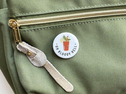 The Oh Bloody Hell Bloody Mary Pinback Button by Onderkast Studio, shown on a green fabric bag with a gold zipper and suede pull, displays a fun cocktail design—ideal for brunch lovers seeking high-quality accessories.