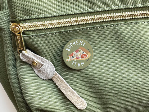 The Supreme Team Pizza Pinback Button by Onderkast Studio, featuring a pizza slice design and SUPREME TEAM text, is attached to a light green canvas bag with gold zipper and suede pull—a must-have for any Pizza Aficionado.