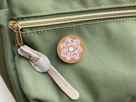 The Sprinkle Donut Pinback Button by Onderkast Studio, featuring a pink-iced donut with colorful sprinkles, is attached to the pocket of a green fabric bag with a gold zipper and suede pull—a perfect accessory for donut lovers.