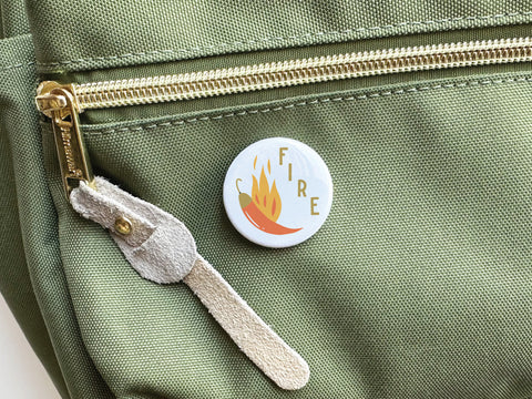 The Fire Chili Pinback Button by Onderkast Studio features bold FIRE text and flame art on a red background, displayed on a light green bag with a gold zipper and suede pull.