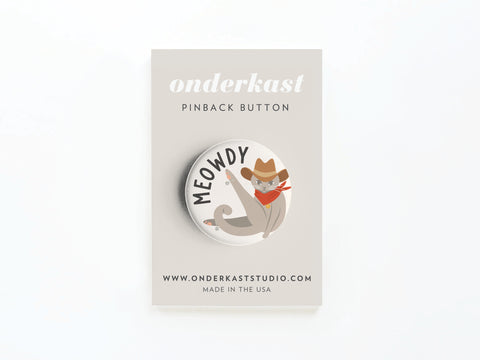 The Meowdy Pinback Button by Onderkast Studio features a cartoon cat in a cowboy hat and red bandana. Printed in the USA, it comes on an Onderkast pinback button card with www.onderkaststudio.com. Perfect for cat lovers!.