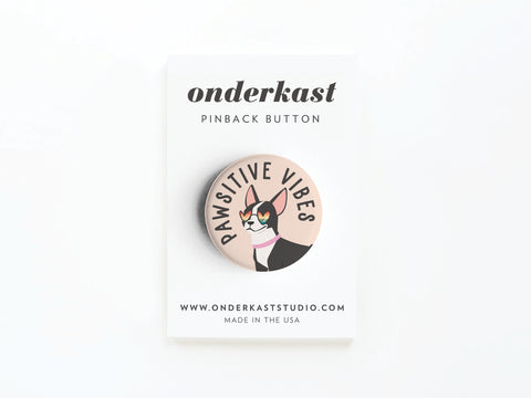 A high-quality pinback button by Onderkast Studio featuring a cool dog and “Pawsitive Vibe,” attached to a card labeled Onderkast PINBACK BUTTON, with the website and “MADE IN THE USA” printed below.