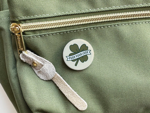The Onderkast Studio Push Your Luck Clover Pinback Button, featuring a four-leaf clover and banner design, adds Irish charm when displayed on a green bag with gold zipper and suede pull.