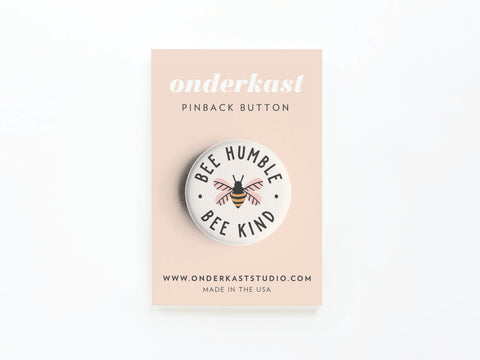 The Bee Humble Bee Kind Pinback Button by Onderkast Studio features a whimsical bee design and inspiring words on a light pink card reading ONDERKAST PINBACK BUTTON, website, and MADE IN THE USA.