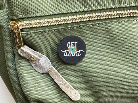 The Get Turnt Vinyl Pinback Button by Onderkast Studio, a black circular button with GET turnt in white and blue, accents the front pocket of a green fabric bag with a gold zipper—perfect for party animals and music lovers.