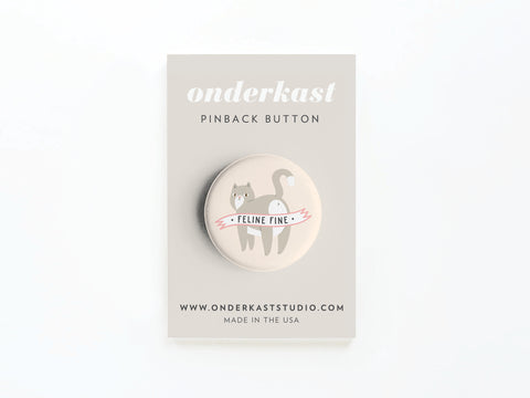 The Feline Fine Cat Pinback Button by Onderkast Studio showcases a smiling gray cat with a “FELINE FINE” banner, attached to a beige Onderkast Studio PINBACK BUTTON card—perfect for any feline fan.