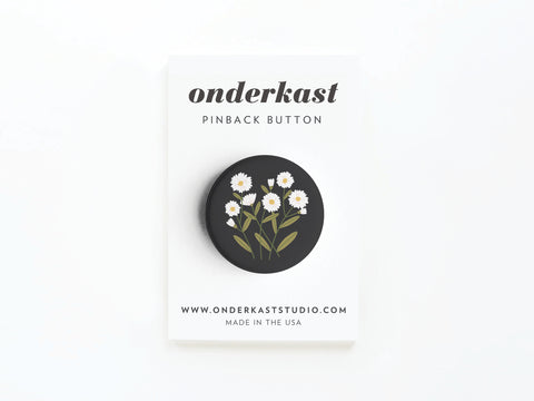 The Daisies Pinback Button by Onderkast Studio is a round black button with white daisies and green leaves, displayed on a card labeled Onderkast Pinback Button, featuring onderkaststudio.com and Made in the USA.