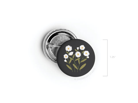 The Daisies Pinback Button by Onderkast Studio features white daisy-like flowers with green stems on a black background and measures 1.25 inches in diameter. Shown open next to its shiny metal back.