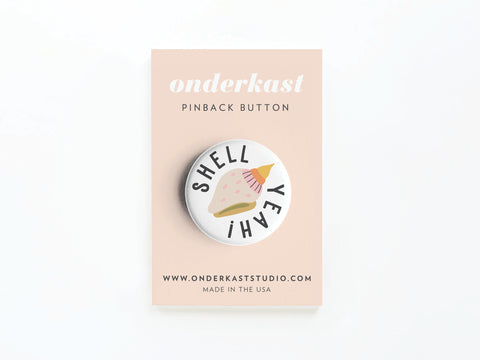 The Shell Yeah Pinback Button by Onderkast Studio features a pink seashell and “SHELL YEAH” on a round button. Displayed on a blush card labeled onderkast PINBACK BUTTON, it’s perfect for ocean fans. MADE IN THE USA appears at the bottom.