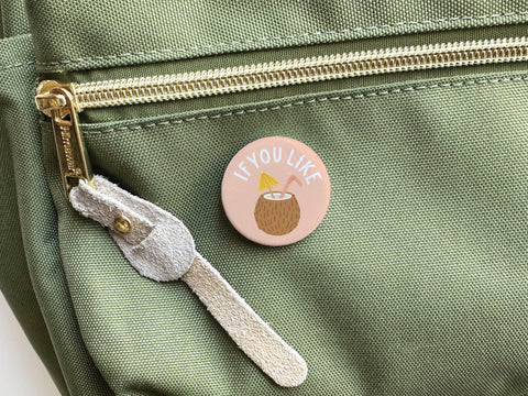 The If You Like Piña Coladas Pinback Button by Onderkast Studio, with a coconut drink design and pink background, is attached to a light green zippered bag—perfect for cocktail lovers or tropical treat fans.