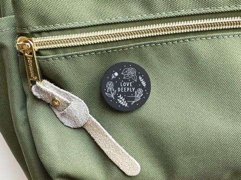 A green fabric bag with gold zipper and suede pull, plus a 1.25 onderkast studio “Love Deeply” pinback button—vivid colors, white floral designs, and LOVE DEEPLY text. Product: Love Deeply Pinback Button – DISCONTINUED.
