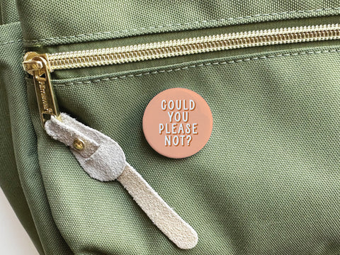 The Onderkast Studio Could You Please Not Pinback Button – DISCONTINUED features printed colors and white text, and is shown attached to a light green fabric bag with a gold zipper and suede pull.