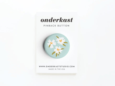 The White Flowers Pinback Button by Onderkast Studio features white and yellow daisies on pastel blue, displayed on a card labeled onderkast pinback button, printed in the USA with onderkaststudio.com at the bottom.