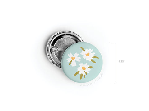 The White Flowers Pinback Button by Onderkast Studio features three white daisies with yellow centers and green leaves on a light blue background. Printed in the USA, this round pin measures 1.25 inches in diameter.