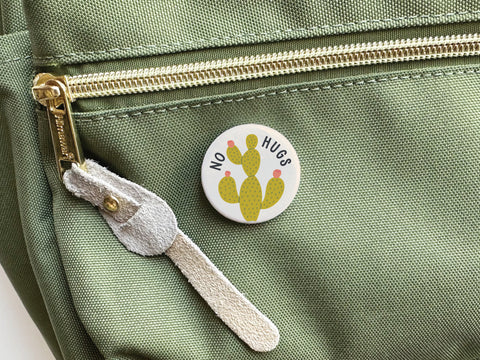 Onderkast Studio’s No Hugs Cactus Pinback Button, featuring a cactus and “NO HUGS,” comes on a green fabric bag with gold zipper and suede pull—perfect for playful introverts who value personal space.