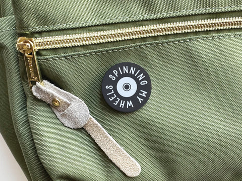 A discontinued Onderkast Studio Spinning My Wheels pinback button, featuring vibrant colors and quality production, is attached to a green fabric bag with a gold zipper and suede pull.