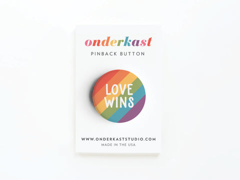 The Love Wins Pinback Button by onderkast studio features rainbow stripes and celebrates LGBTQ+ rights. Displayed on a white card with onderkaststudio.com and Made in the USA—perfect for allies and activists.