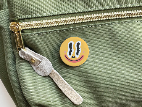 The Onderkast Studio Trippy Happy Face Pinback Button—a round yellow pin with an abstract, wavy black and white design—is attached to a green backpack near a gold zipper with a tan suede pull.