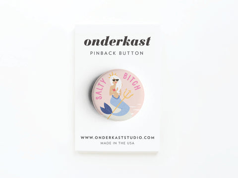 The Salty Bitch Mermaid Pinback Button by Onderkast Studio features a mermaid with sunglasses and trident, plus “SALTY BITCH”—perfect for ocean lovers. Attached to a card labeled “onderkast pinback button.”.